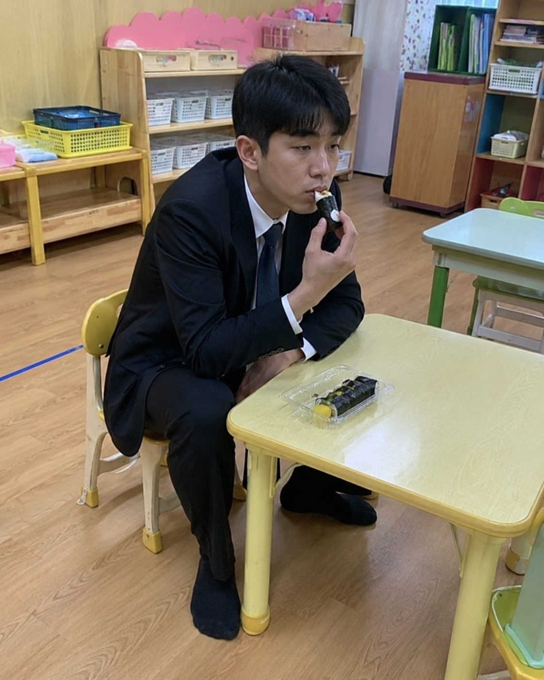 어린이집 교실의 노란 작은 책상과 의자에 정장을 입은 청년이 앉아 김밥을 집어 먹는 모습