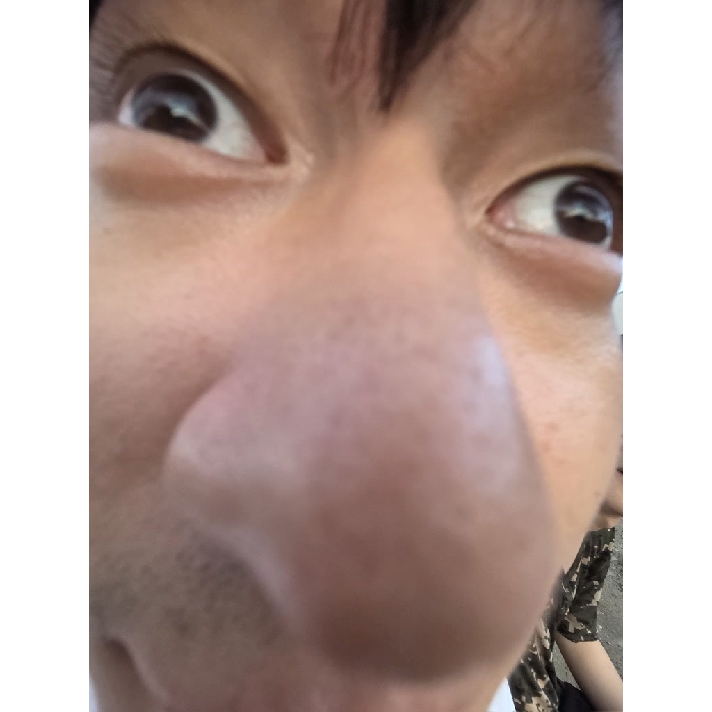 카메라를 아주 가까이 대고 찍어 코가 화면 중앙을 가득 채운 사람의 얼굴 클로즈업. 두 눈은 옆쪽을 바라보고 있고 오른쪽 아래에 카무플라주 무늬 소매가 걸쳐 있다.